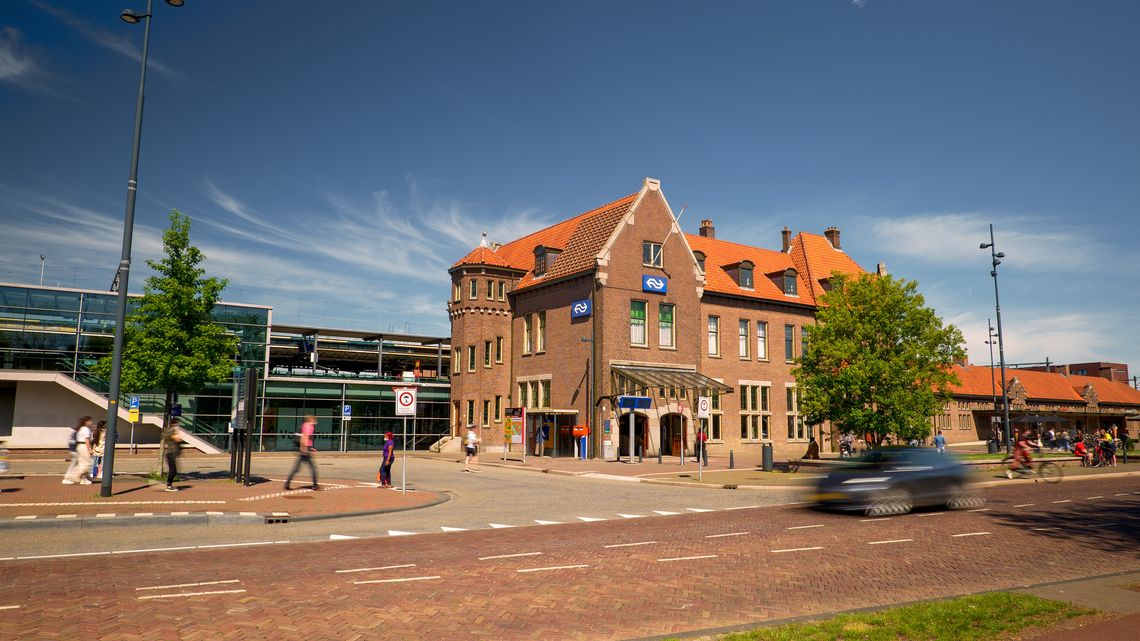 Het station van Deventer aan de voorzijde van de stationshal. We zien mensen voor het station langs lopen en er rijdt een auto.